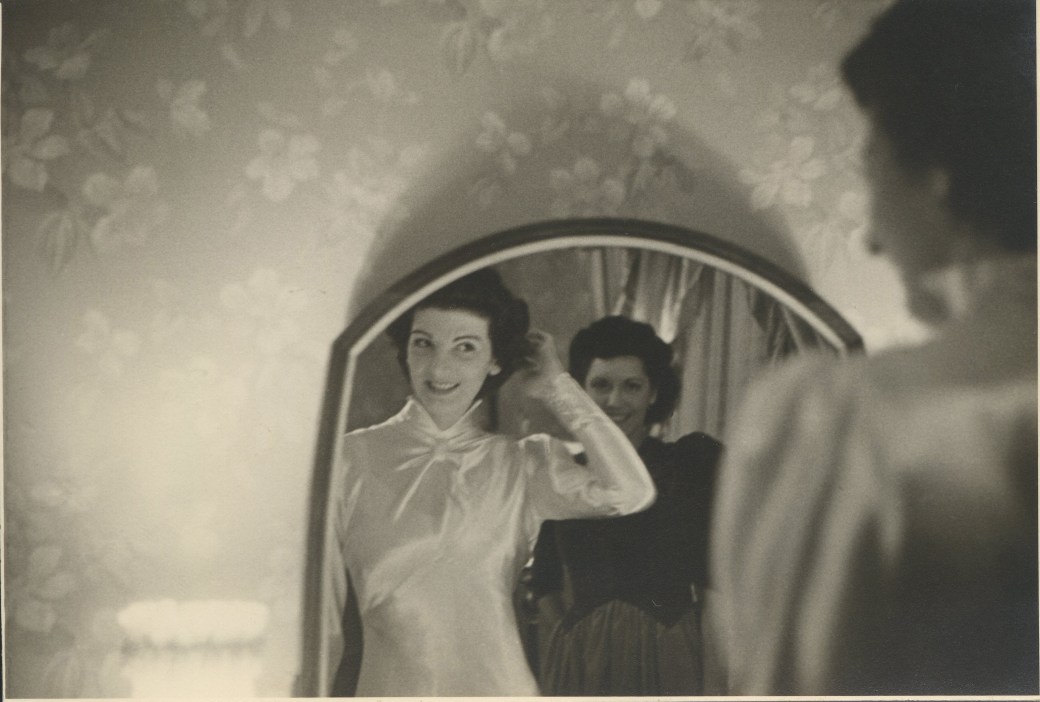 Two women looking into a mirror, one wearing a white dress.
